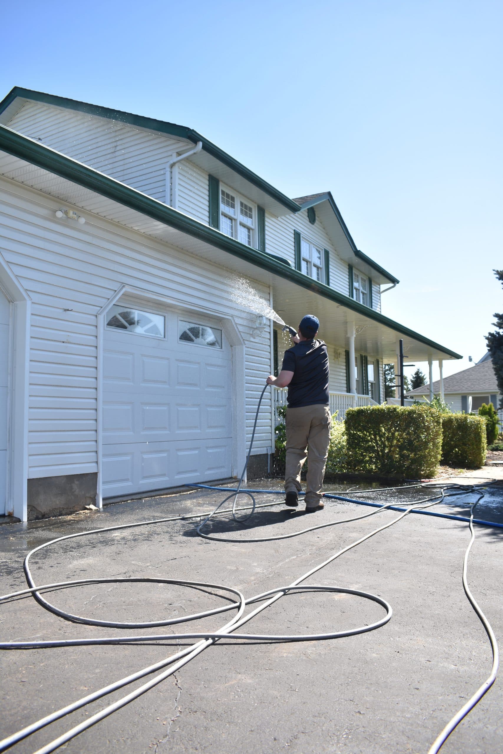 A Gladiator Pro Wash technician soft-washing a white two-storey farmhouse, supply hoses coiled across the driveway