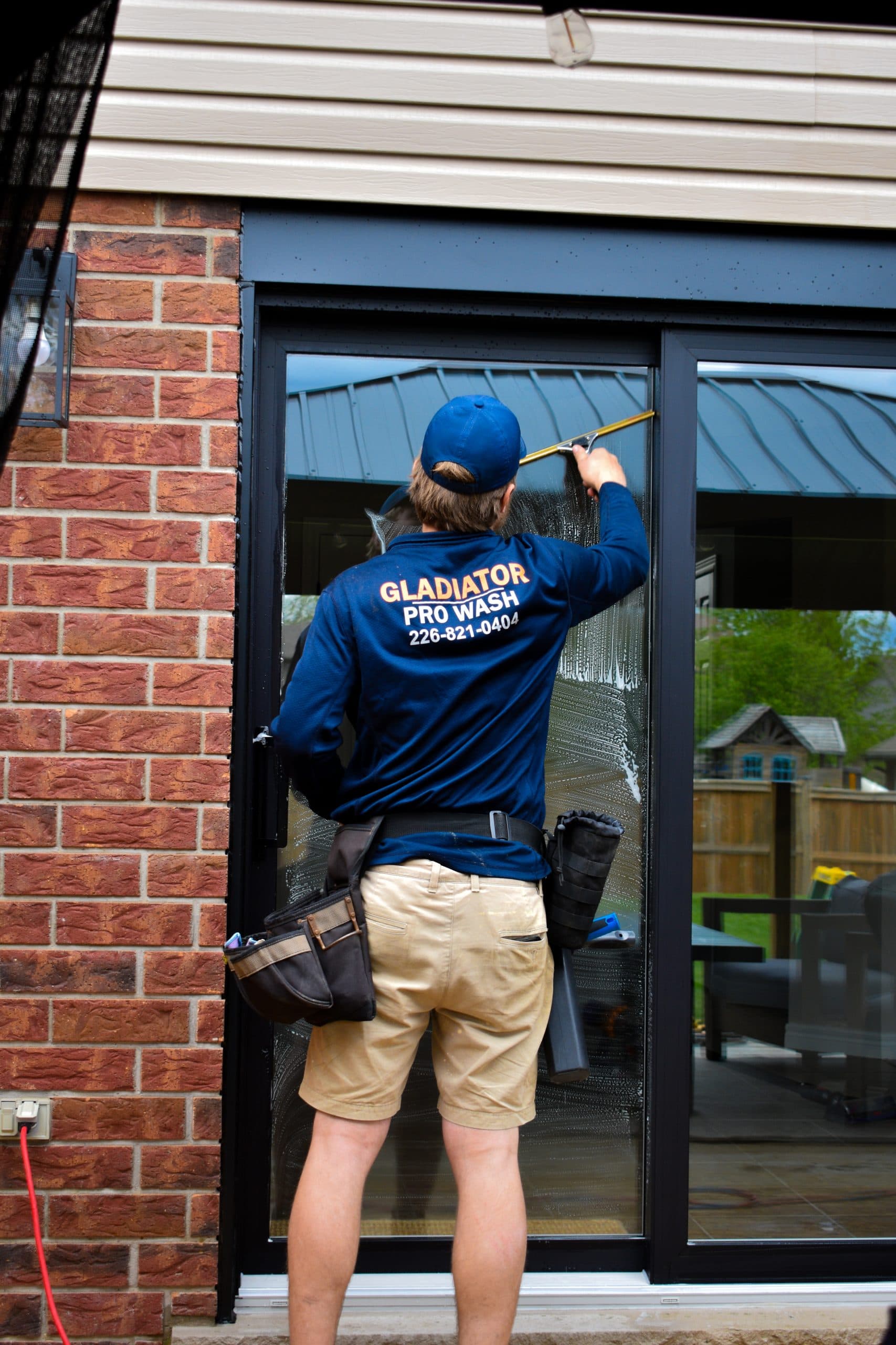 A technician in a Gladiator Pro Wash shirt squeegeeing a patio door beside red brick