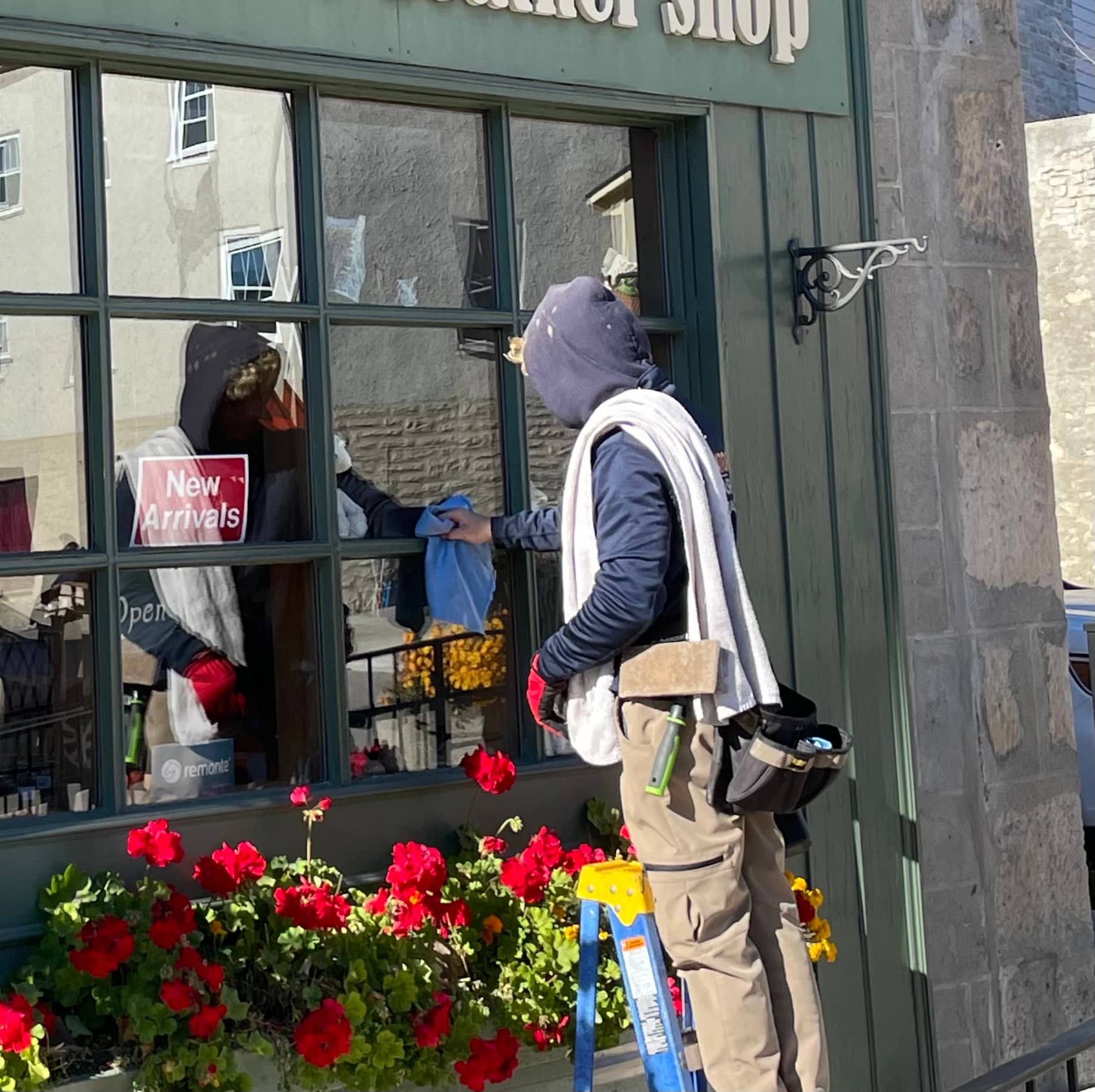 A technician cleaning a small-town shop window above a planter of red geraniums