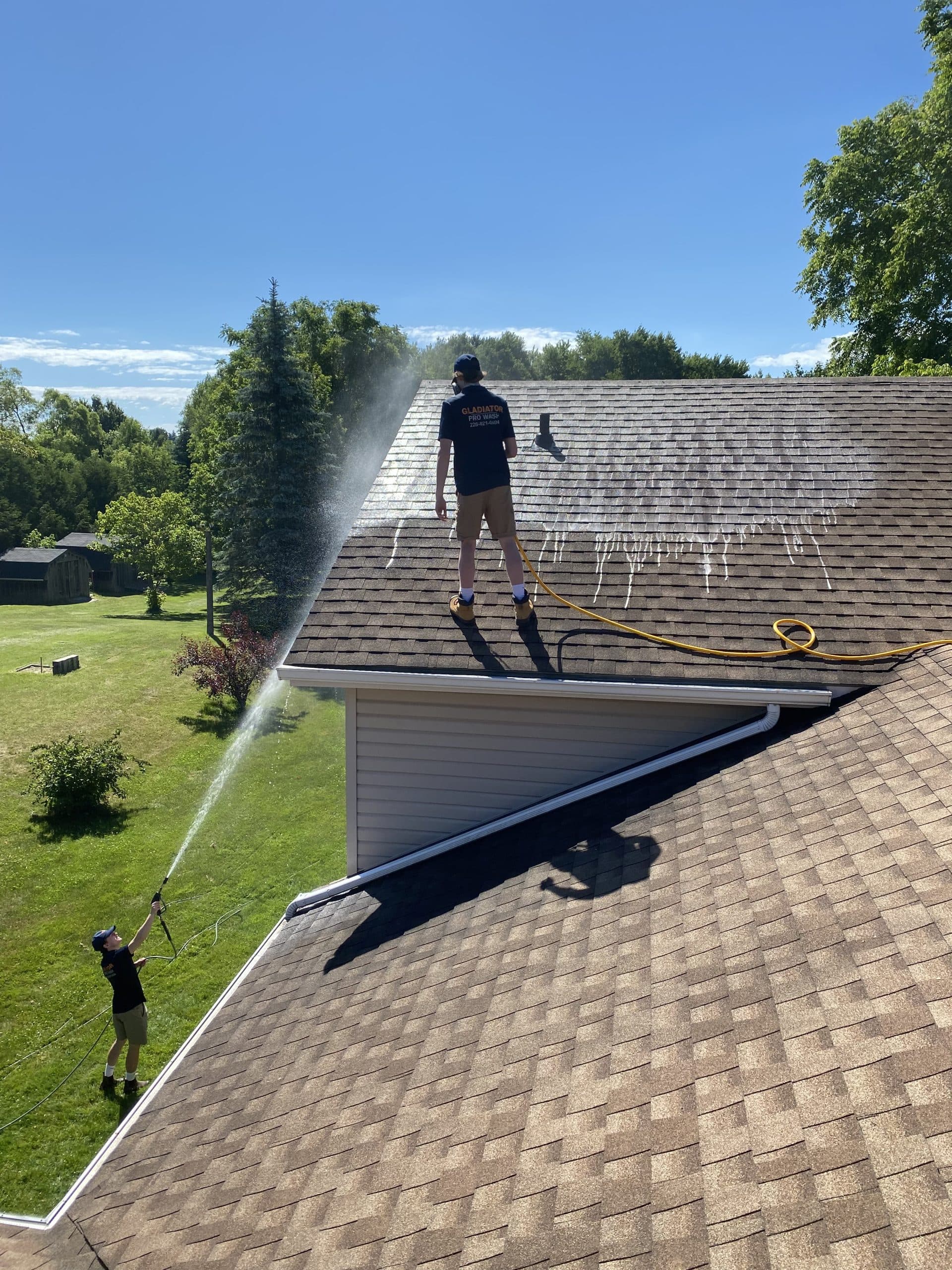 Two technicians on a shingled roof, a water arc catching the sun against a blue summer sky